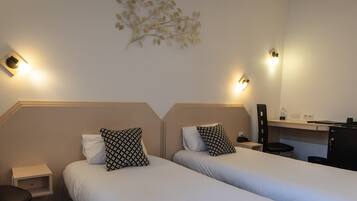 A hotel room with two beds, each with patterned pillows, a small nightstand, a desk with a chair, and wall-mounted lights.