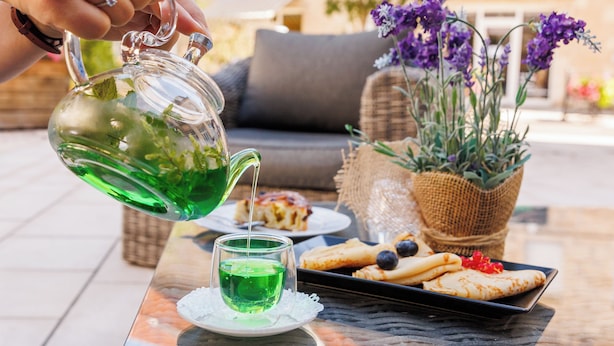 Eine Person gießt grünen Tee in ein Glas auf einem Tisch. Es gibt einen Korbstuhl, eine Auswahl an Gebäck und einen Blumentopf mit violetten Blüten.