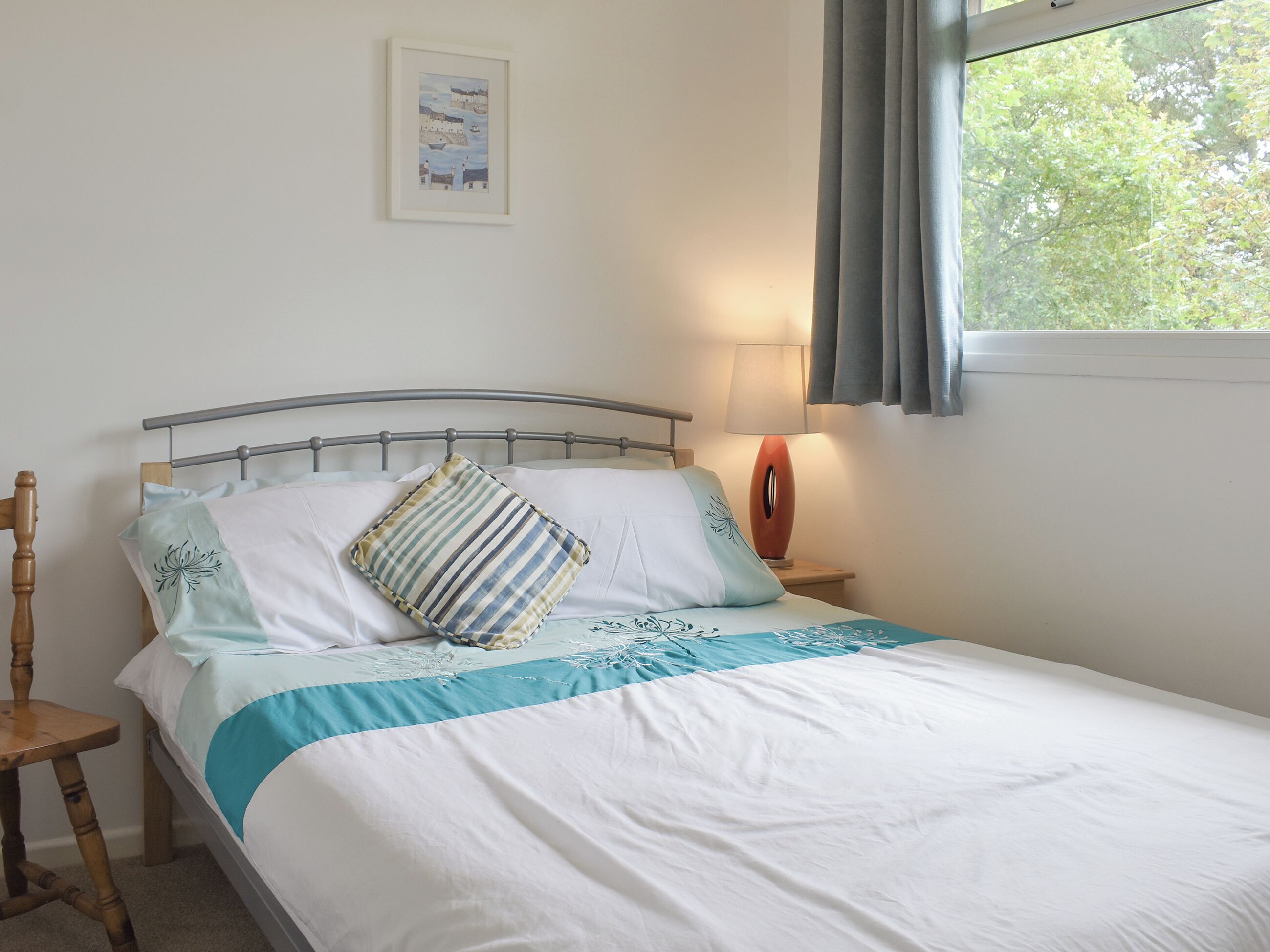 Una cama con estructura metálica, una lámpara de noche, un cuadro en la pared y una ventana con cortinas.