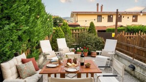 Une terrasse avec une table en bois dressée pour le repas, entourée de chaises blanches et d’un canapé.