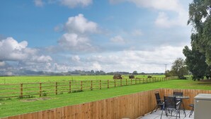 Un patio con vistas a un campo verde y fardos de heno, cerca de una valla de madera y mobiliario exterior.