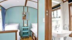 A compact, well-lit room with a wooden ceiling, a small teal bedside table with a mirror, two beds with white linens, and a marble bathroom with a sink and window. - Brunel Boutique Railway Carriage 5 (Dawlish Warren)