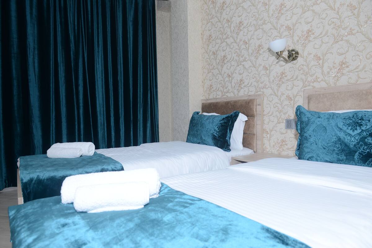 A hotel room with two beds, each with a blue bedspread and white linens. There is a wall-mounted light fixture above the beds.