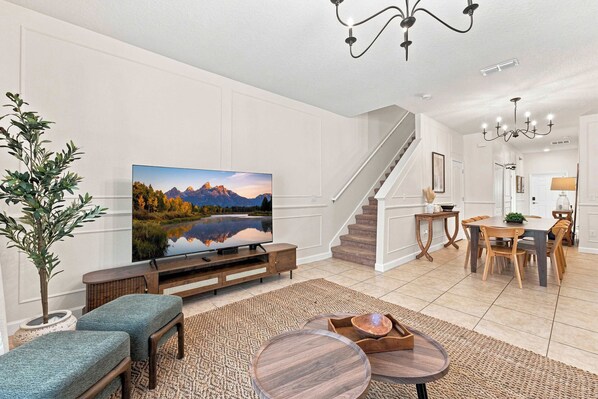 A modern living room with a flat-screen TV, a staircase, a dining area, and a large plant.