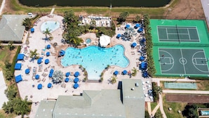 An aerial view of a resort with a large swimming pool, tennis courts, and sun loungers.