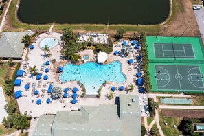 An aerial view of a resort with a large swimming pool, tennis courts, and sun loungers.