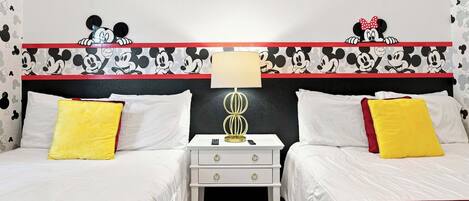 Habitación de hotel con dos camas, una mesita de noche con lámpara y papel tapiz con temática de Mickey Mouse.