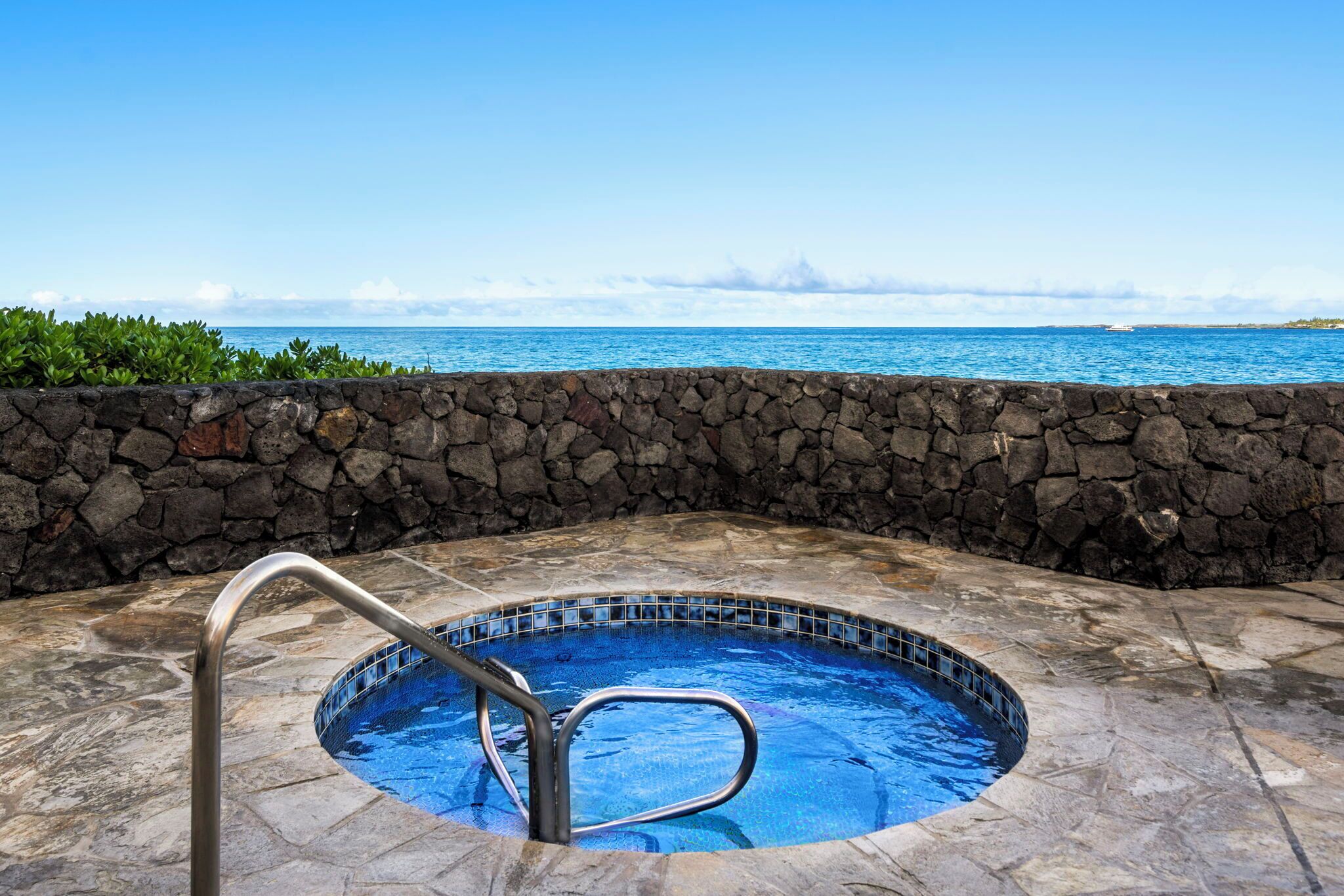 A stone-walled hot tub with a metal ladder, surrounded by stone tiles, overlooking a clear blue ocean.