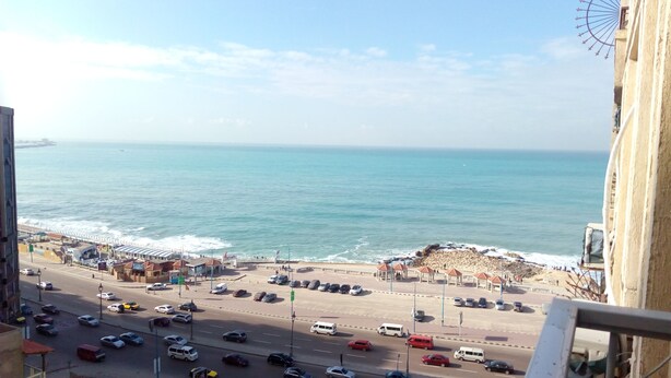 Un paisaje urbano costero con una playa, una carretera con coches y una noria al fondo.