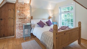A bedroom with a sloped ceiling, a brick wall, a wooden bed, a nightstand, and a window with a view of greenery. - The Coach House Stables (Hernhill)