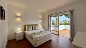 A bedroom with a bed, bedside tables, a view through a sliding glass door, and a white wall.
