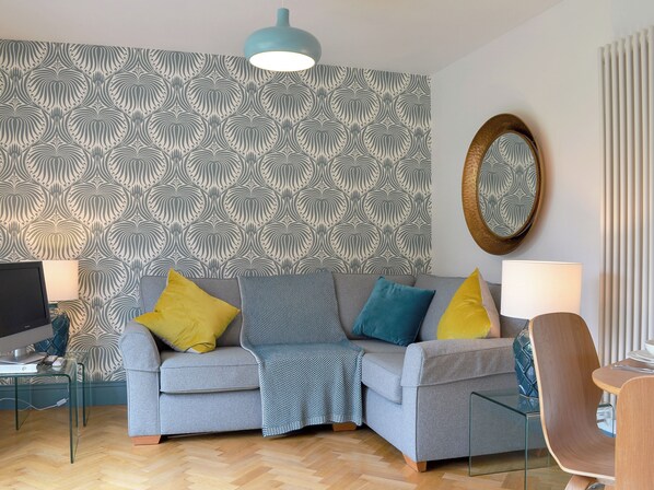 Una sala de estar moderna con un sofá gris, cojines amarillos y azules, una mesa de centro de vidrio, un televisor y un papel tapiz estampado.