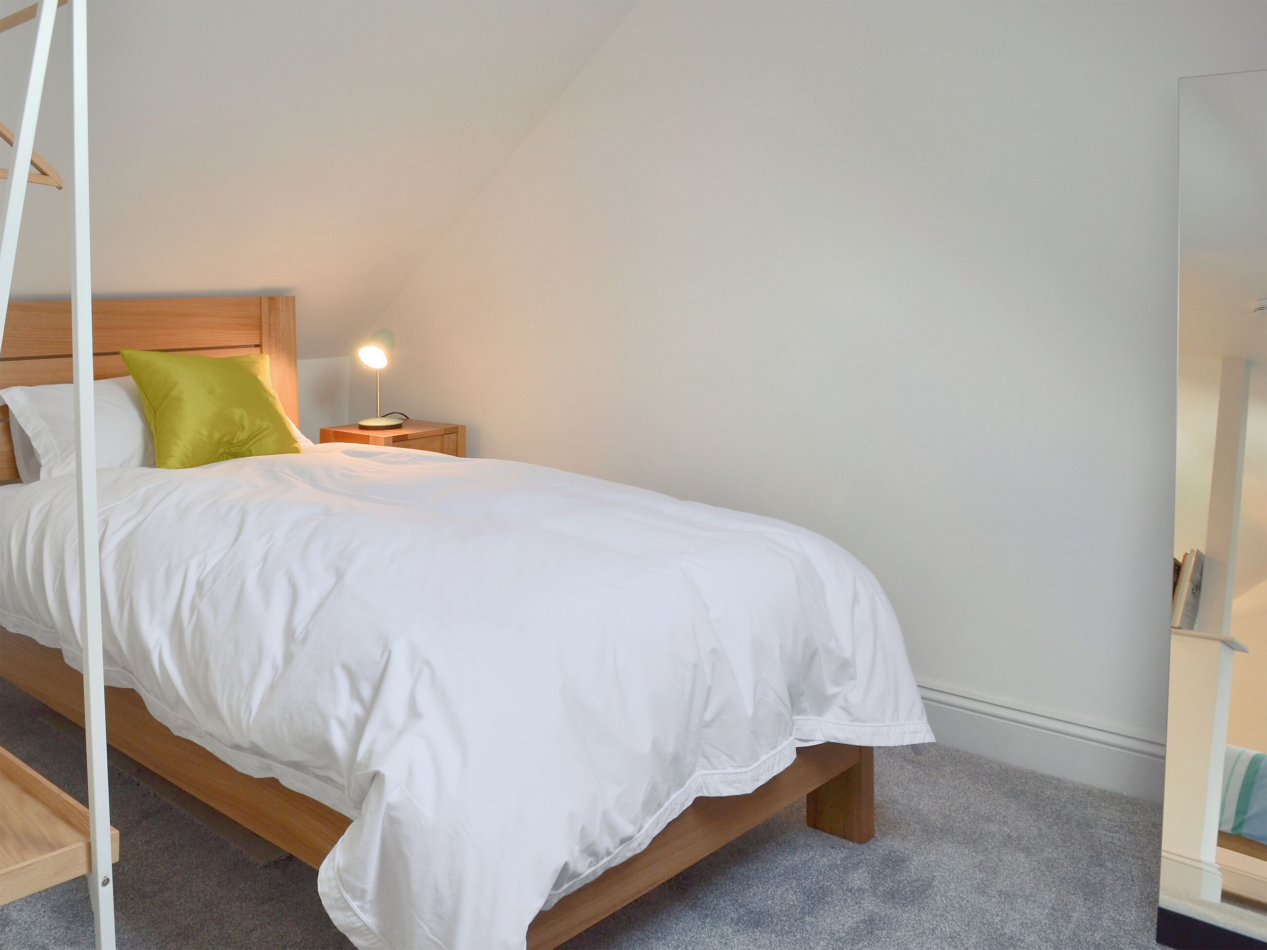 Un lit bien fait, avec une literie blanche et un oreiller vert, une lampe de chevet et une tête de lit en bois, dans une pièce dont le plafond est en pente.