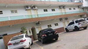A multi-story building with a parking area. There are several cars parked in front, including a white car and a black car. The building has multiple air conditioning units and windows.