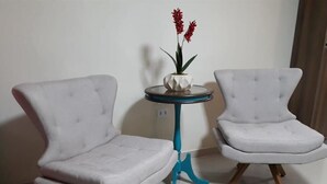 Two white upholstered chairs with wooden legs and a small round table with a blue base and a white vase with red flowers.