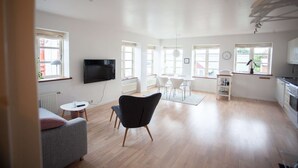 A spacious living area with a flat-screen TV, a dining table with chairs, a kitchen area, and large windows allowing natural light.