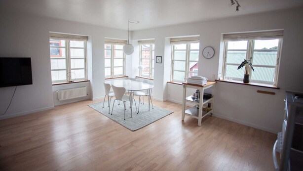 A spacious room with a wooden floor, a dining table with chairs, a TV mounted on the wall, and large windows providing natural light.