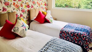 Una habitación con dos camas, papel tapiz floral y una ventana con vista a follaje.