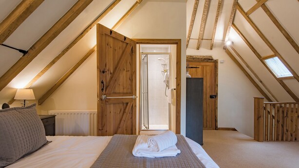 A bedroom with a sloped ceiling, wooden beams, a bed with a quilted bedspread, a bedside table with a lamp, and a door leading to a bathroom. - 3 bedroom accommodation in Hollingbourne (Hollingbourne)