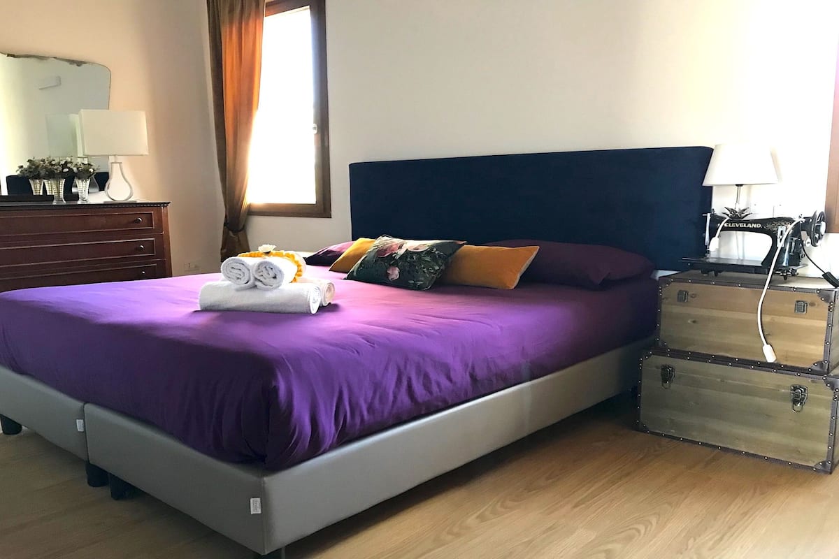 Un lit bien fait, avec une literie violette, une commode avec un miroir et une valise posée sur le sol.