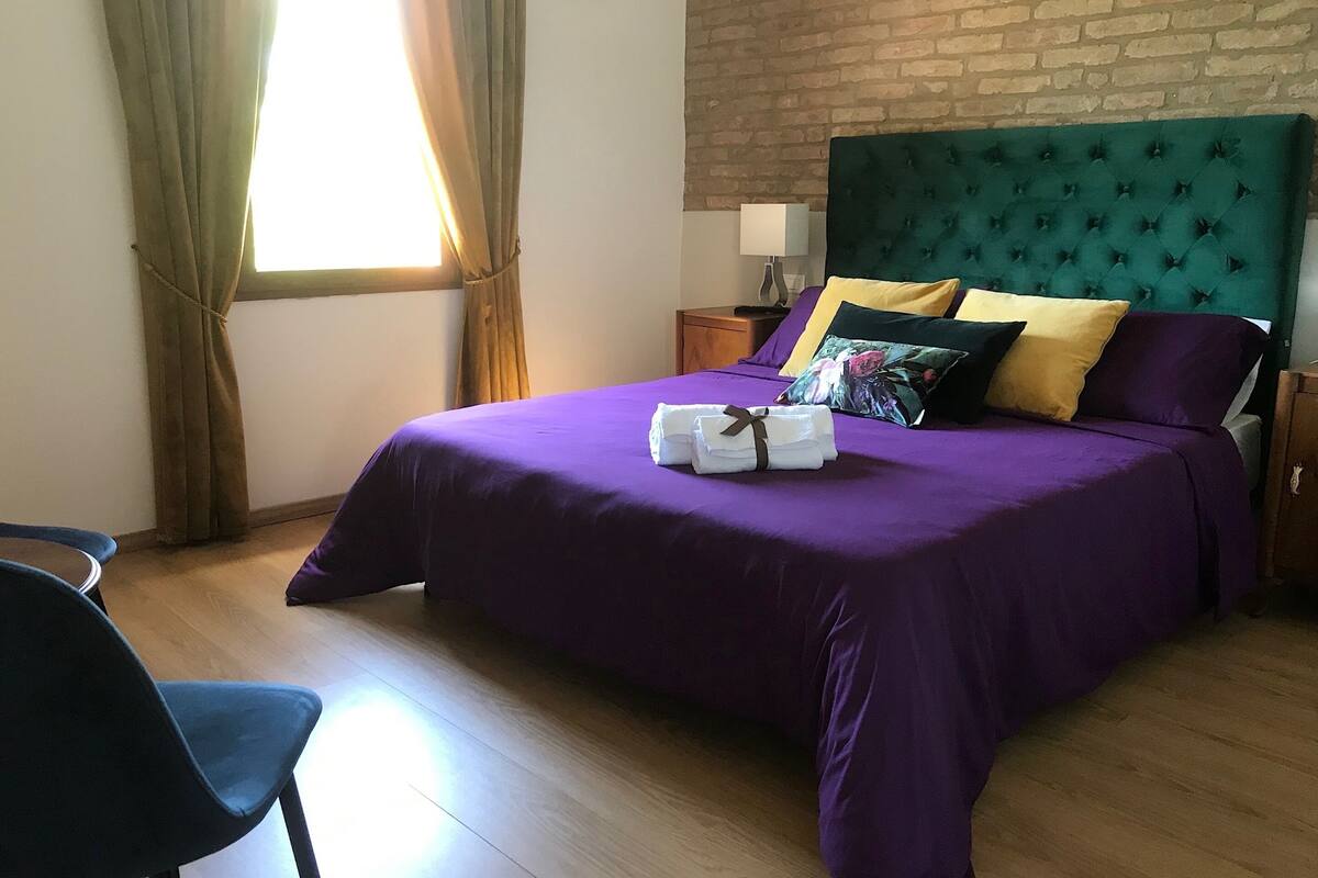 Un lit bien fait, recouvert d’une couvre-lit violette et agrémenté d’oreillers, une table de chevet en bois avec une lampe, et une chaise dans une pièce dont un mur est en briques et qui possède une fenêtre avec des rideaux.