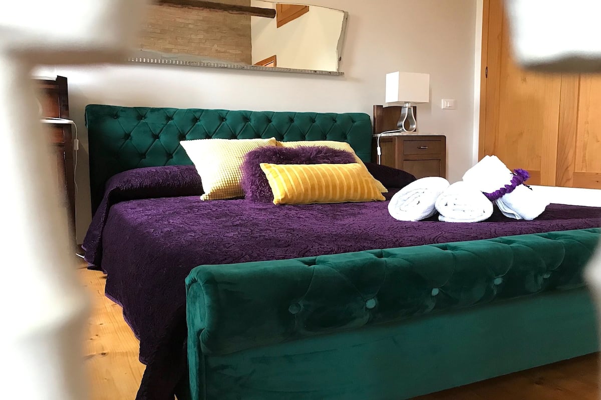 Un lit avec une tête de lit verte, des coussins violets et jaunes, et un banc recouvert d’un tissu vert.