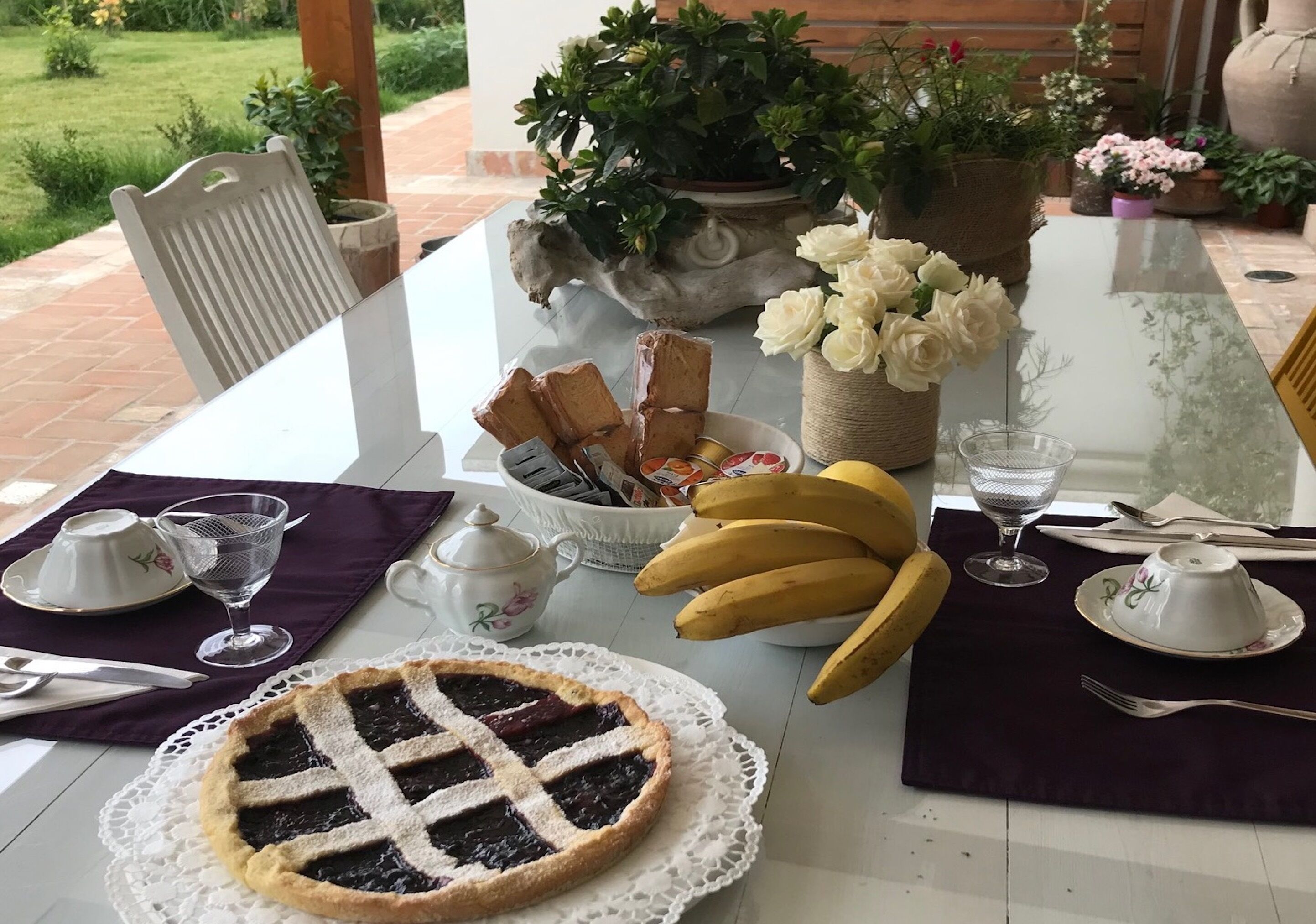 A table set with a pie, bananas, a basket of bread, and a vase of flowers.