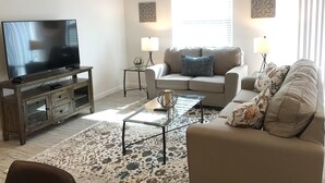 A living room with a flat-screen TV, a wooden entertainment center, a beige sofa, a glass coffee table, and a patterned rug. - CHARMING 2 BDRM 2 BATH Sm Dog Friendly Patio Villa w/FREE WIFI (The Villages)