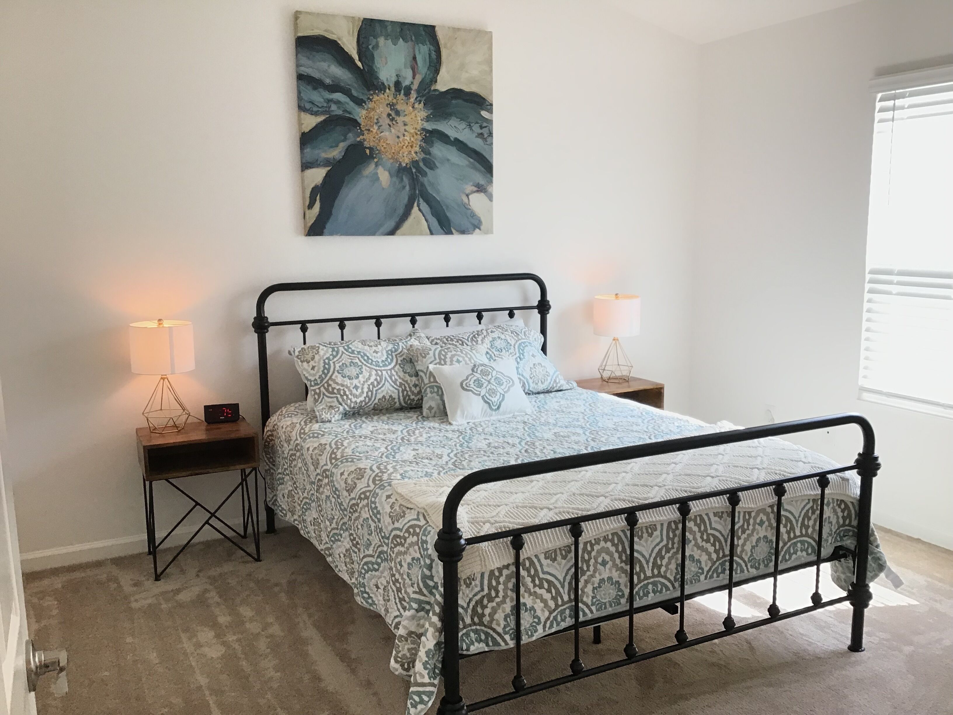 Un dormitorio con estructura de cama metálica, ropa de cama estampada, dos lámparas de noche, un reloj de pared y un cuadro floral grande.