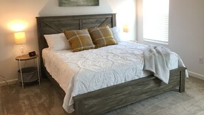 A bedroom with a bed, a nightstand with a lamp, a painting of a house, and a window. - CHARMING 2 BDRM 2 BATH Sm Dog Friendly Patio Villa w/FREE WIFI (The Villages)