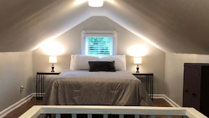 Ein Schlafzimmer mit schrägen Wänden, zwei Nachttischlampen, einem Fenster mit Jalousien, einem Bett mit grauer Bettdecke und einem Holzboden.