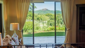 Ein Zimmer mit großem Fenster, Vorhängen, Deckenventilator und Blick auf eine grüne Landschaft mit Bäumen und Hügel.