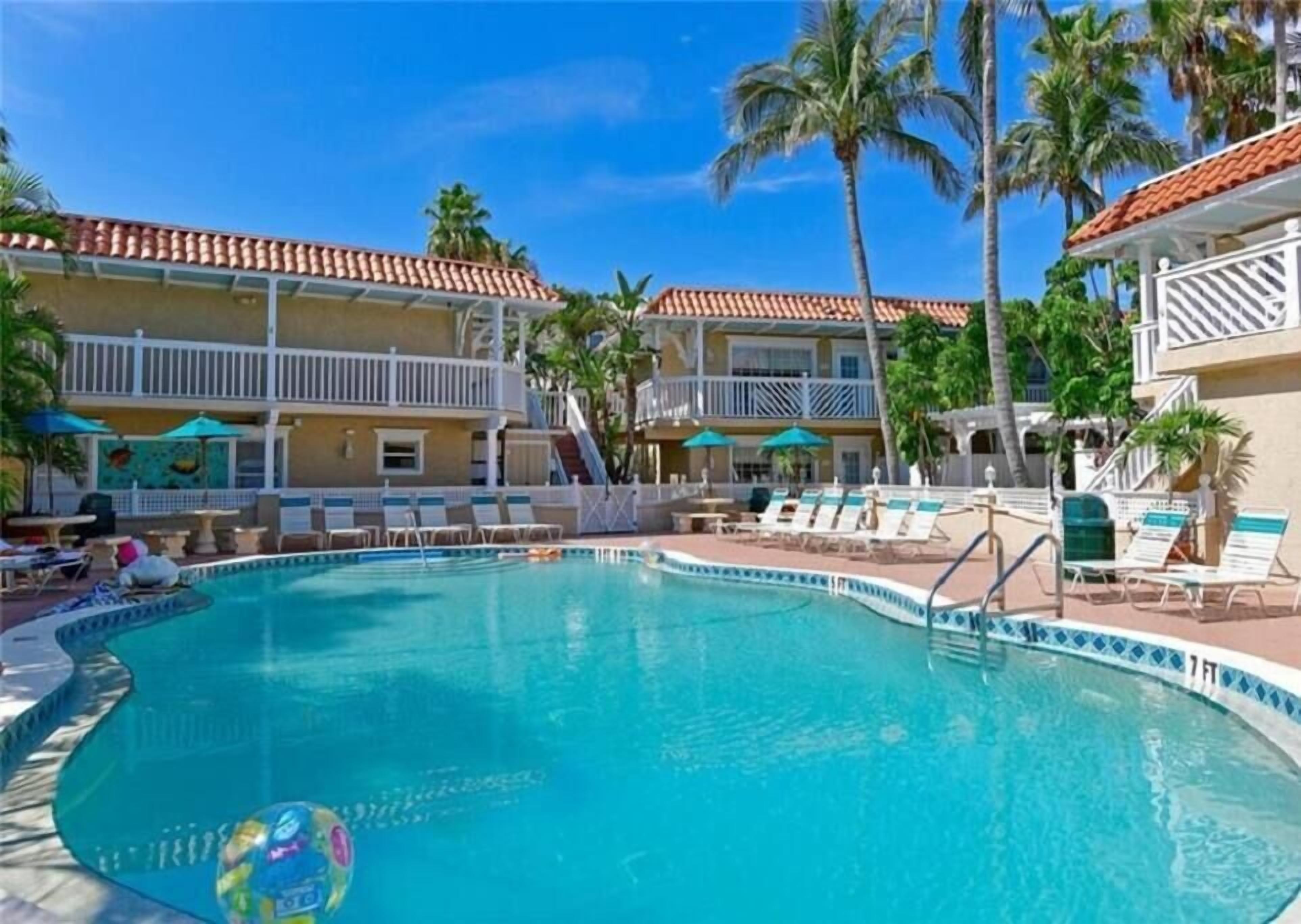 A swimming pool with lounge chairs and umbrellas, surrounded by palm trees and a building with a tiled roof.