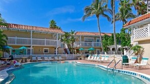 A swimming pool with lounge chairs and umbrellas, surrounded by palm trees and a building with a tiled roof. - Tortuga Inn Beach Resort # 229 Island Condo, 2Bdrm (Bradenton Beach)