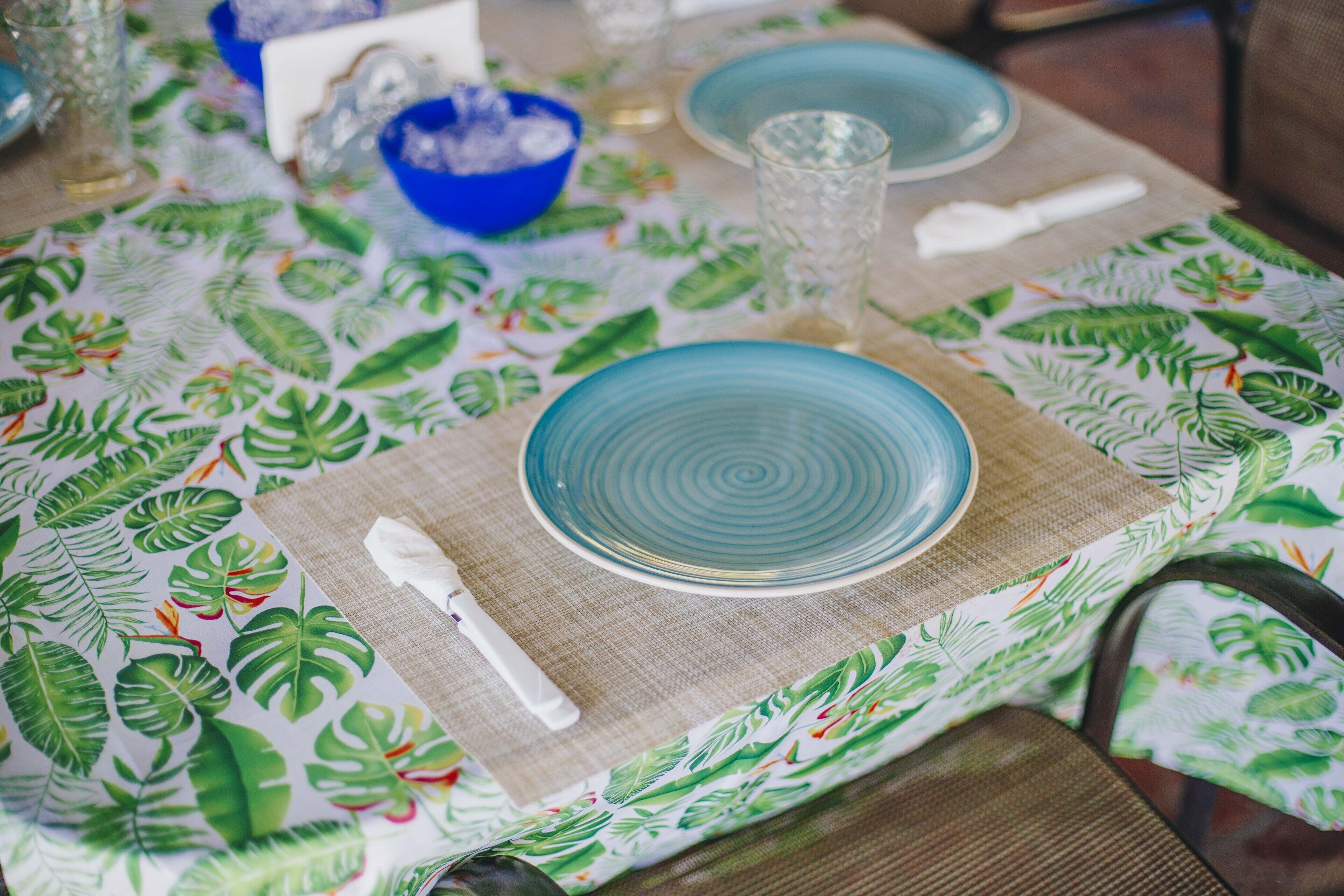 A table set with blue plates, a clear glass, and a white napkin on a tablecloth with a tropical leaf pattern.
