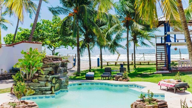 A pool with a stone waterfall, palm trees, beach view, lounge chairs, and a small structure with a ladder.