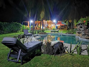 Un espace piscine avec des chaises longues, une structure en pierre et un bâtiment dont les fenêtres sont illuminées en arrière-plan.
