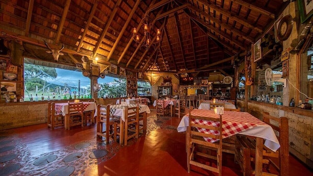 Een rustiek restaurant met houten tafels en stoelen, een groot raam met uitzicht op een schilderachtige omgeving, en een houten plafond met zichtbare balken.