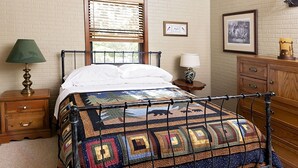 A bedroom with a metal bed frame, a ceiling fan, a window with blinds, a wooden dresser, and a nightstand with a lamp.