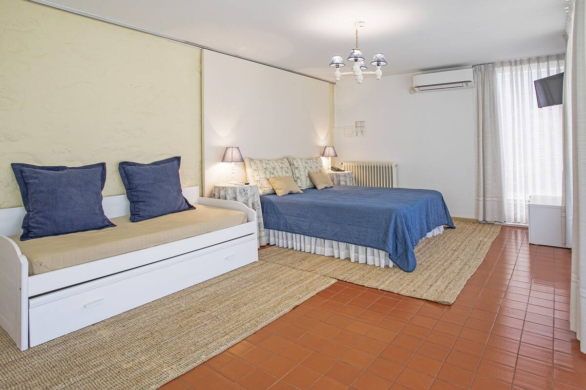 Une chambre avec un lit blanc, un canapé blanc, une petite table de chevet avec des lampes, un radiateur et un téléviseur fixé au mur.