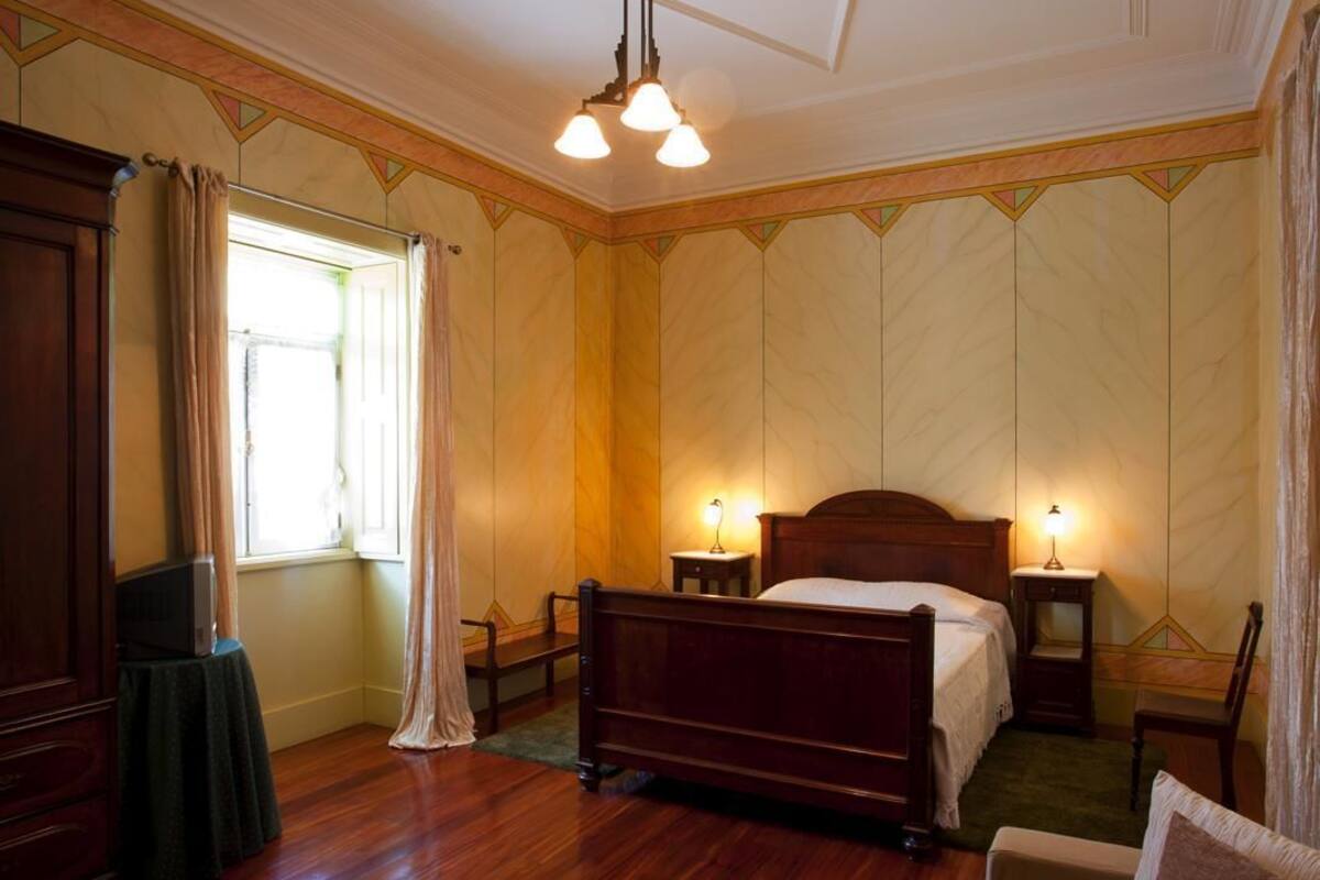 Une chambre à coucher avec un lit en bois, deux tables de chevet avec des lampes, une fenêtre avec des rideaux et une petite table sur laquelle se trouve un téléviseur.