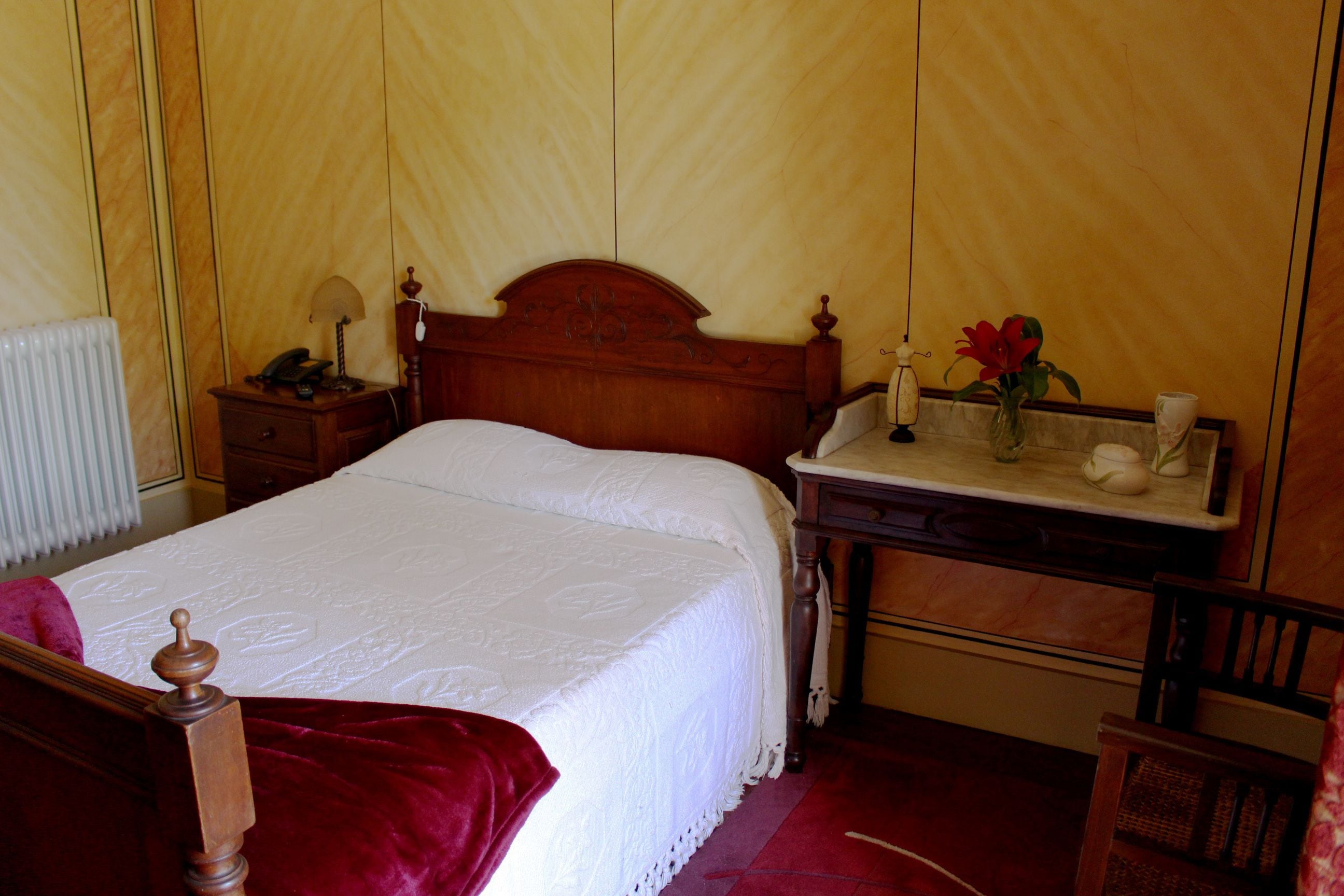 Una cama bien hecha con cabecera de madera, una mesita de noche con lámpara y un florero con una flor.