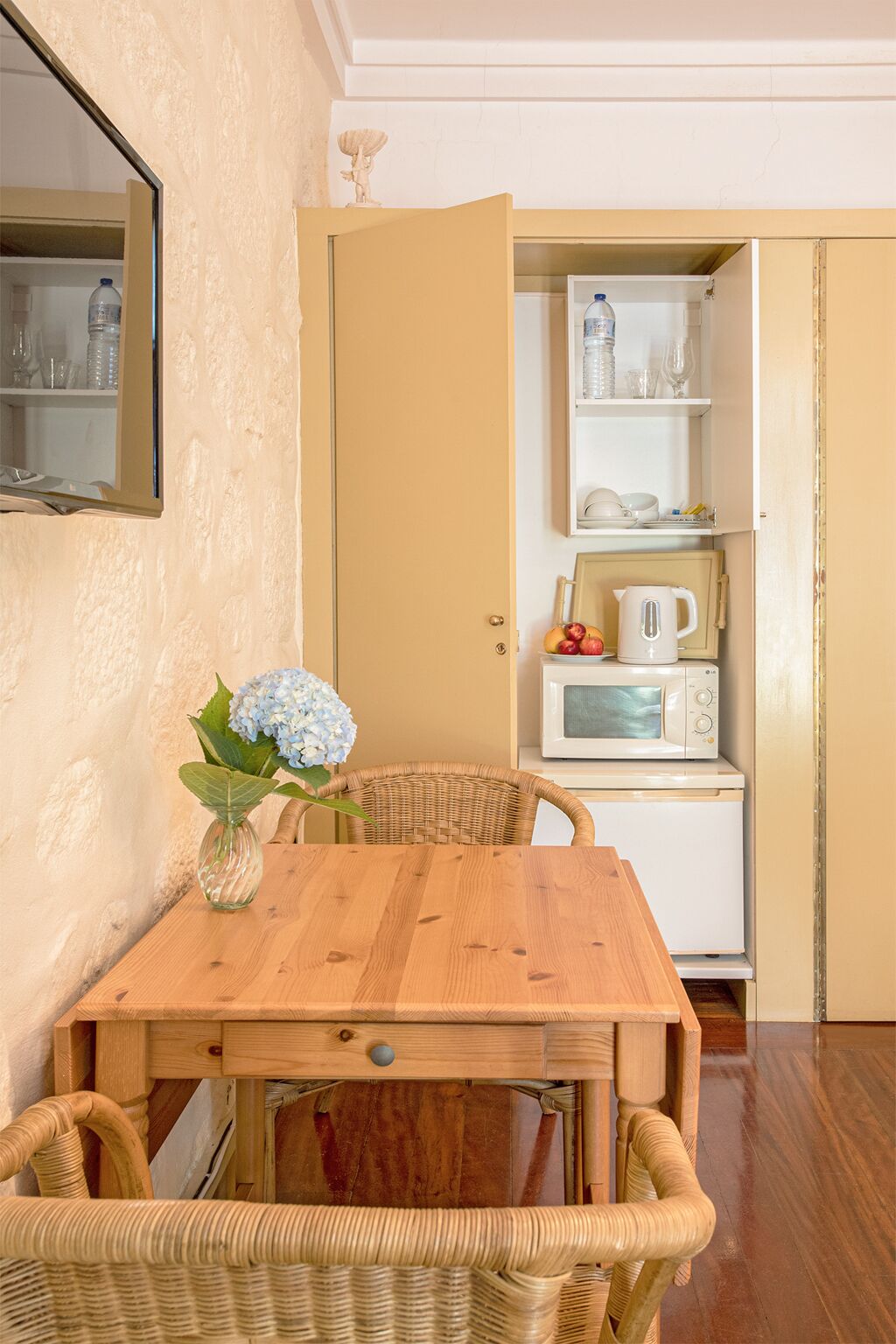 Una mesa de comedor de madera con sillas de mimbre, un florero y una zona de cocina con microondas, hervidor y estanterías.