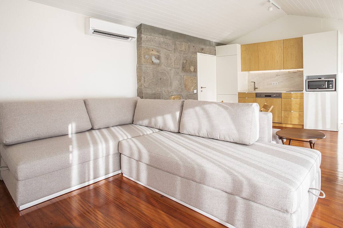 Un salon moderne doté d’un canapé d’angle, d’un mur d’accent en pierre, d’un coin cuisine avec des meubles en bois et d’un système de climatisation intégré.