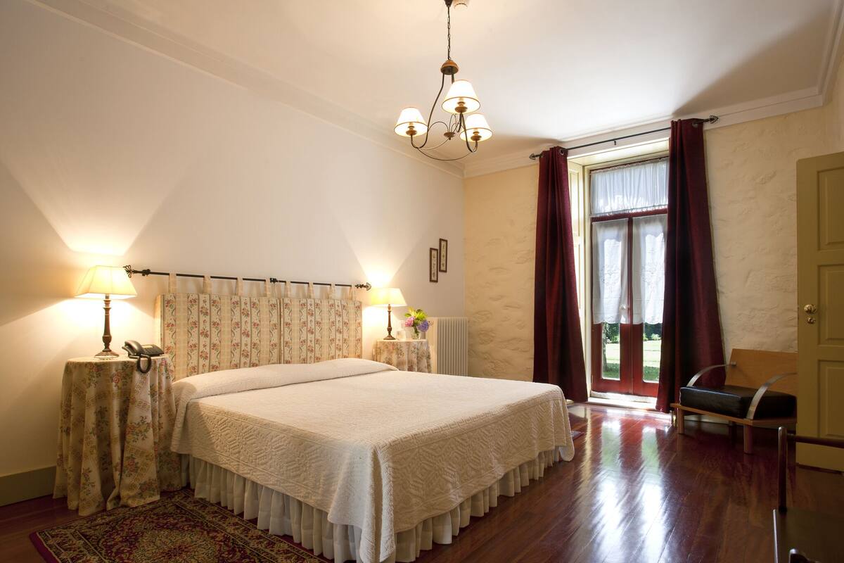 Une chambre spacieuse avec un grand lit, un lustre, un plancher en bois et une vue sur l’extérieur, visible à travers une fenêtre ornée de rideaux.