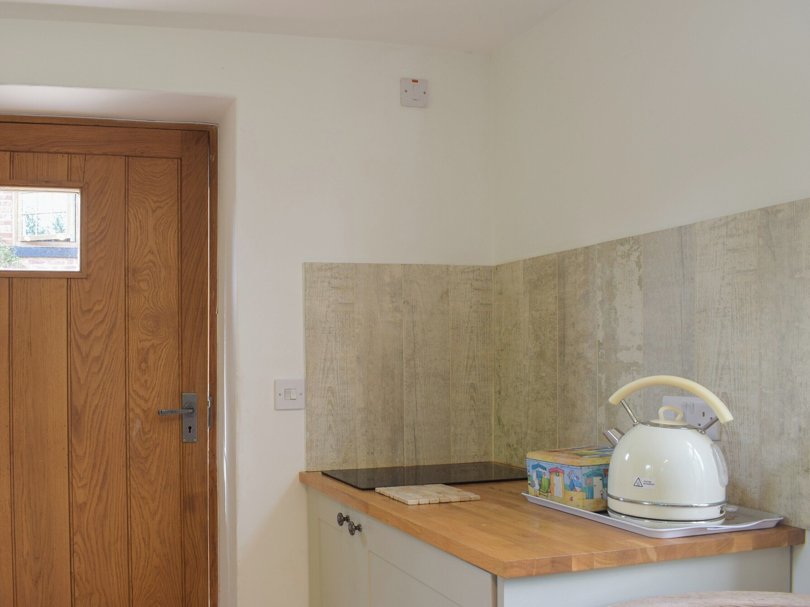 Una cocina con una puerta de madera, una pared blanca con un interruptor de luz y una tetera blanca sobre la encimera.