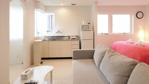 A modern kitchen with white cabinets, a sink, and a microwave. There is a small table with a remote control and a box. A gray sofa with a red blanket is in the living area, and a clock is on the wall. - Kukuru House (Naha)