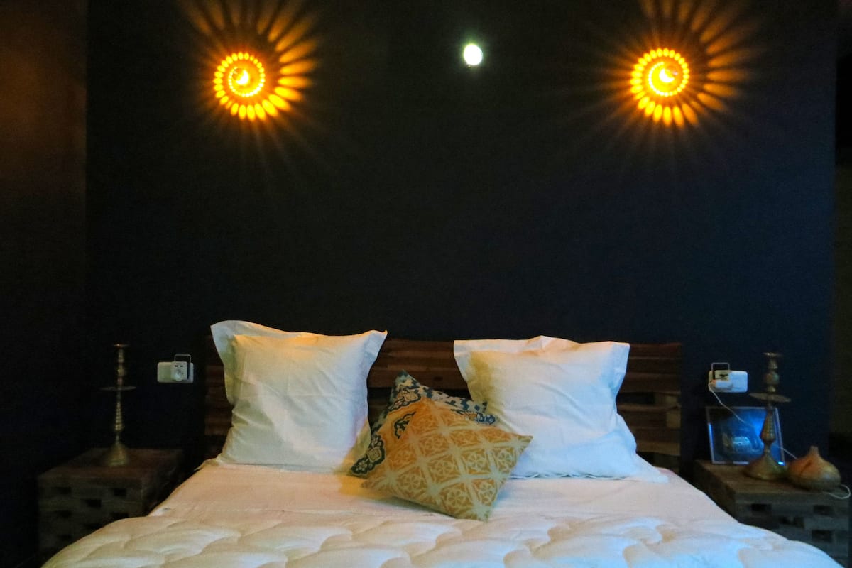 Uma cama com roupa de cama branca, duas almofadas e uma almofada decorativa. Há duas luminárias fixadas na parede com design semelhante ao sol.
