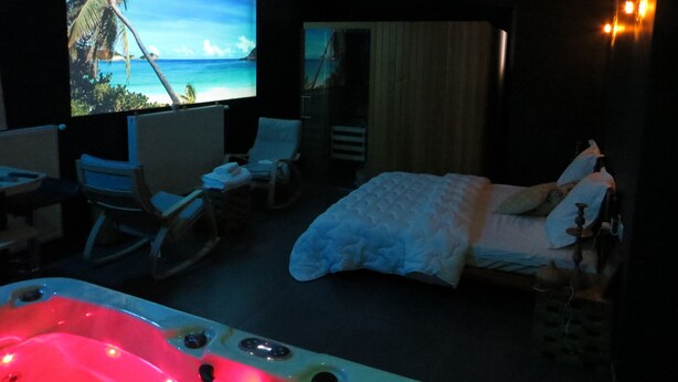 Une chambre d’hôtel avec un grand écran de télévision diffusant une scène de plage, un lit, une baignoire avec éclairage, une chaise et une petite table.