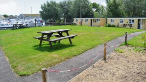 Een gazon met picknicktafels, een pad en een rij boten afgemeerd bij een gebouw met ramen en een deur.